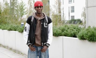 Yacqub Mohamed seisoo pihalla reppu selässä ja hymyilee. Taustalla näkyy kerrostaloja ja vihreää kasvillisuutta.