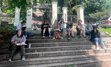 Joukko opiskelijoita istuu vanhoilla rappusilla ulkona. Heillä on piirustusvihot kädessä. Opiskelijat ovat iloisia.