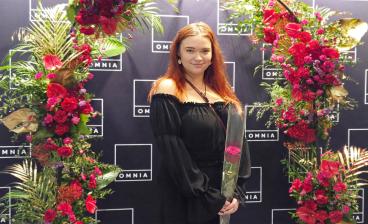 Ronja Oksanen seisoo kukkakaaren alla ruusu kädessään ja hymyilee.