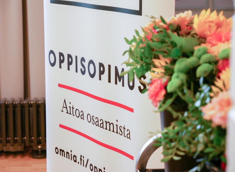 Taustalla roll-up, jossa teksti Oppisopimus, aitoa osaamista. Etualalla kukkia.