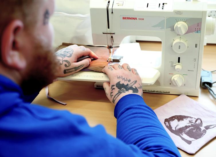 Parrakas henkilö, jolla on paljon tatuointeja käsissään, laittaa kankaan ompelukoneeseen.