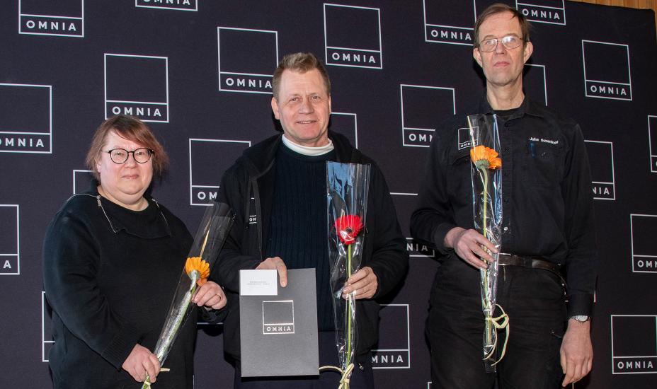 Kolme ihmistä katsoo kameraan käsissään kukat ja kunniakirja.