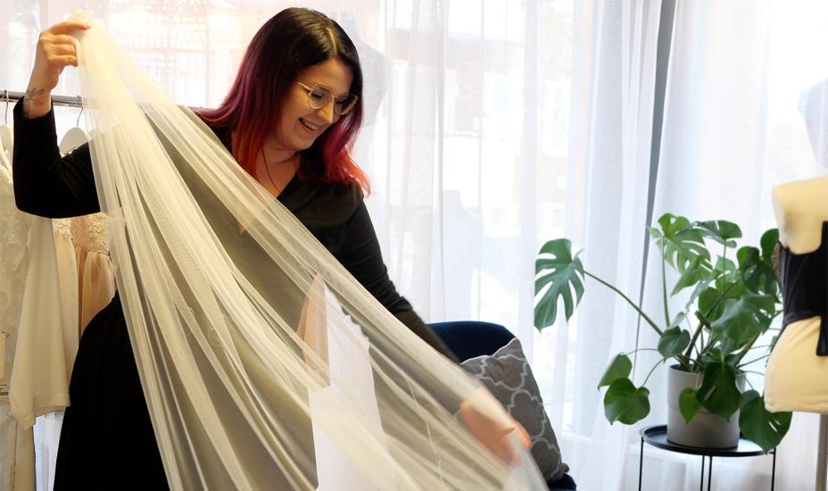 Henkilö pitää kädessään hääpuvun vaaleaa huntua ja levittää sitä. Hän hymyilee.