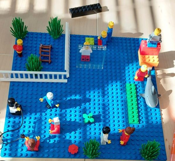 Legorakennelma, jossa on erilaisia legohahmoja. Rakennelmassa osa hahmoista on sisällä ja osa ulkona. Erilaiset tilat on kuvattu legokasveilla ja tekemällä legopalikoista seiniä. Etualalla yksi legohahmo pitää kädessään mikrofonia ja keskustelee kolmen hahmon kanssa.