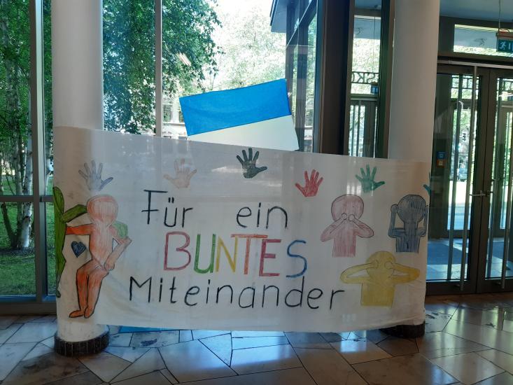 Koulun aulaan ripustettu lakana, jossa on värikkäitä kuvia ja seuraava saksankielinen teksti: Für ein buntes miteinander.