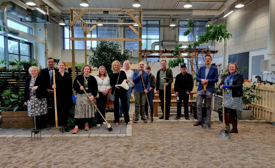 Opetushallituksen kutsumat vieraat Omnian vihersalissa. Maassa on hiekkaa ja taustalla näkyy kasveja sekä puisia puutarharakennelmia. Osalla ihmisistä on käsissään puutarhatyökaluja.