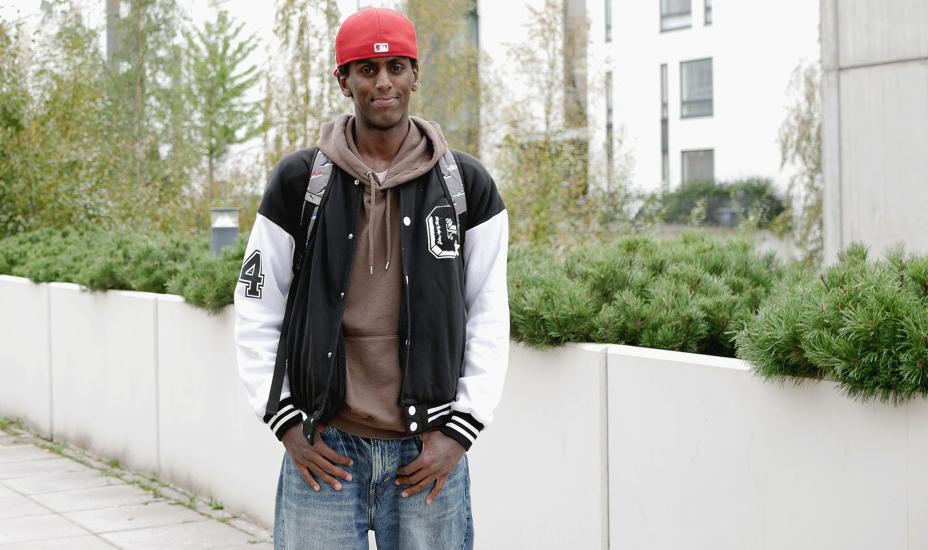Yacqub Mohamed seisoo pihalla reppu selässä ja hymyilee. Taustalla näkyy kerrostaloja ja vihreää kasvillisuutta.