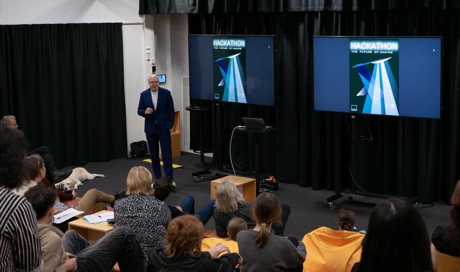 Puhuja pitää hackathonin avauspuhetta kahden tapahtumajulistetta näyttävän näytön välissä, yleisön istuessa lattialla ja tuoleilla; palveluskoira lepää puhujan edessä.