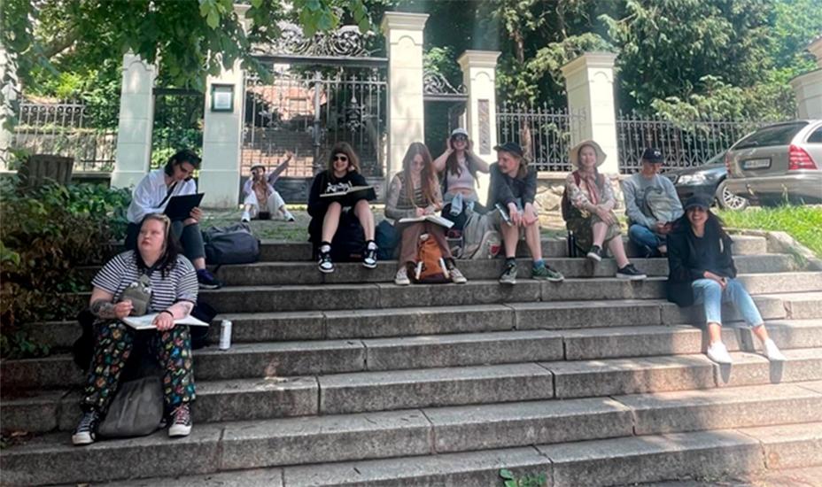 Joukko opiskelijoita istuu vanhoilla rappusilla ulkona. Heillä on piirustusvihot kädessä. Opiskelijat ovat iloisia.