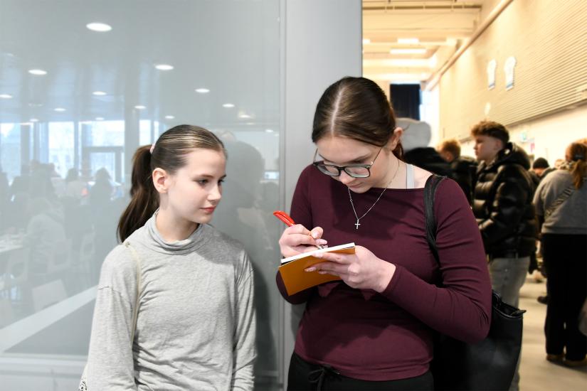 Kaksi nuorta seisoo ihmisporukan edessä. Toisella on kädessään muistilehtiö, johon hän kirjoittaa.