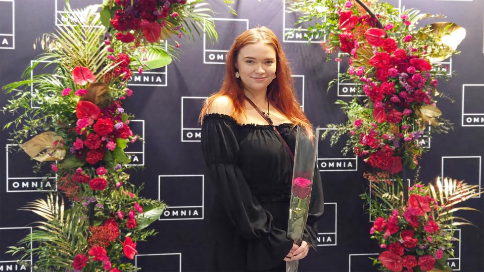 Ronja Oksanen seisoo kukkakaaren alla ruusu kädessään ja hymyilee.