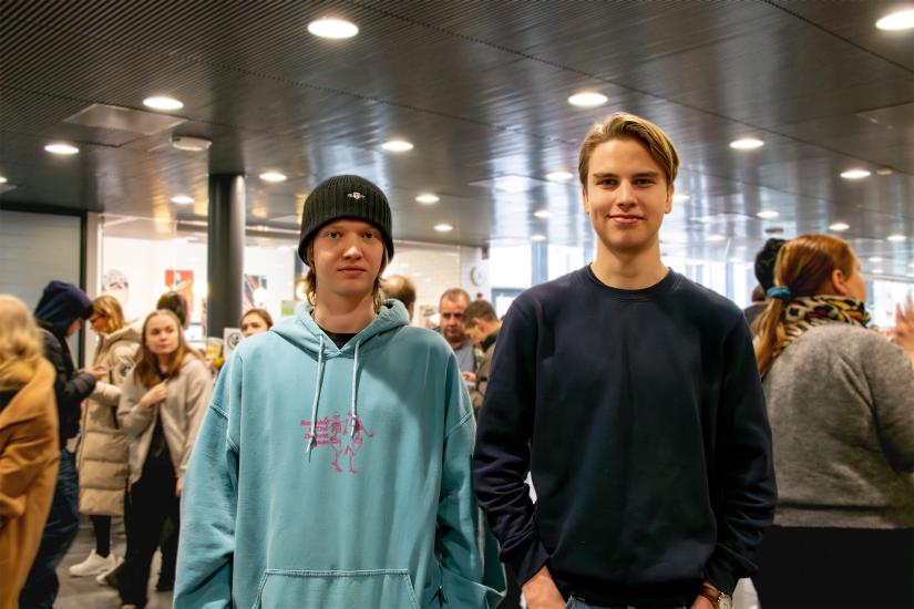 Kaksi nuorta ihmistä seisoo kameran edessä ja katsoo kameraan. Taustalla näkyy ihmisiä.