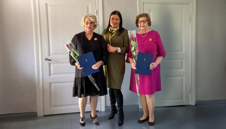 Kolme henkilöä seisoo vierekkäin. Kahdella on kädessään ruusu ja kunniamerkki.