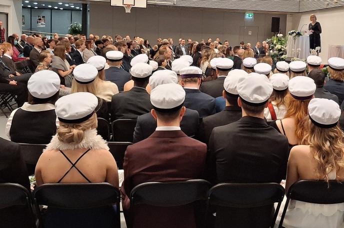 Joukko ylioppilaita istuu liikuntasalissa ylioppilaslakit päässä selkä kameraan päin. Taustalla näkyy puheenpitäjä ja muita ihmisiä.