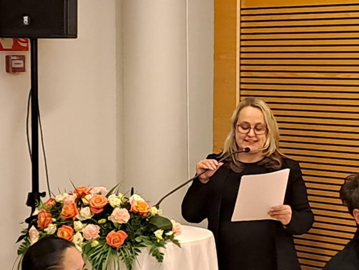 Tanja Rontu-Hokkanen puhuu mikrofoniin. Edessä on pöytä, jossa on juhlavia kukkia..