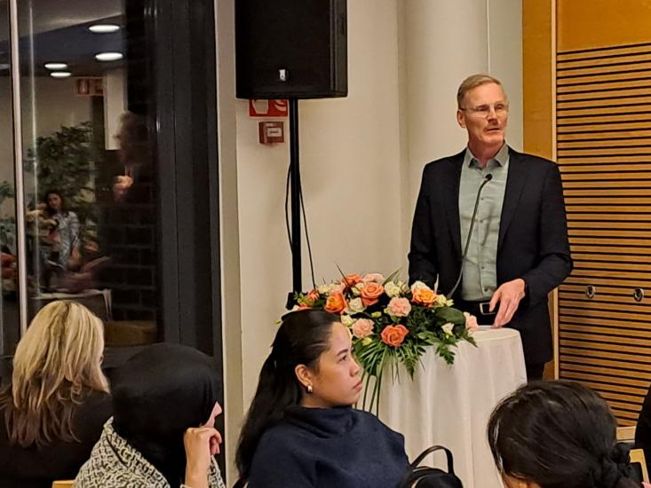 Mies seisoo puhujapöntön takana pitäen puhetta mikrofoniin sisätilassa. Pöytää koristaa kukka-asetelma, ja edessä istuu kuulijoita keskittyneesti seuraamassa esitystä. Taustalla näkyy ikkunan takana lisää ihmisiä ja kasveja.