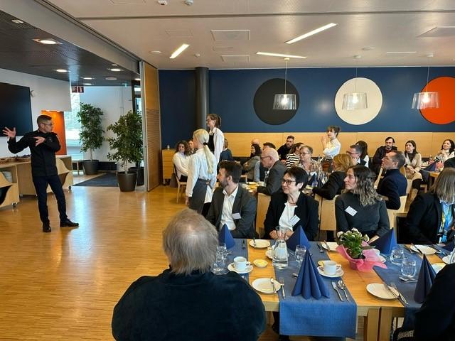 Joukko ihmisiä istuu ruokapöydissä ravintolaympäristössä. Yksi ihminen puhuu edessä.