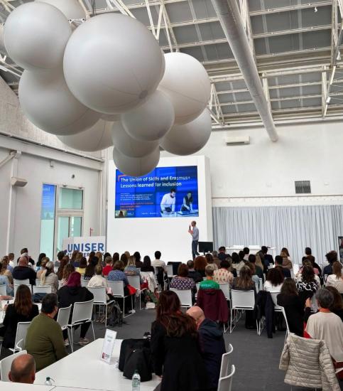 Iso luentosali täynnä osallistujia seuraamassa puhujaa lavalla. Näytöllä näkyy esitys “The Union of Skills and Erasmus+: Lessons learned for inclusion”. Tilassa on moderni vaalea sisustus ja katossa suuria valkoisia ilmapallomaisia muotoja. Tapahtuma järjestetään UNISERin tiloissa.