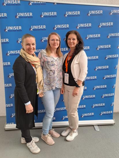 Kolme hymyilevää naista seisoo yhdessä UNISER Teacher Week -taustaseinän edessä. Keskellä seisova henkilö on pukeutunut vaaleaan kukkakuosiseen paitaan ja silmälaseihin. Osallistujilla on kaulanauhat ja nimikyltit, ja tunnelma on ystävällinen ja rento.