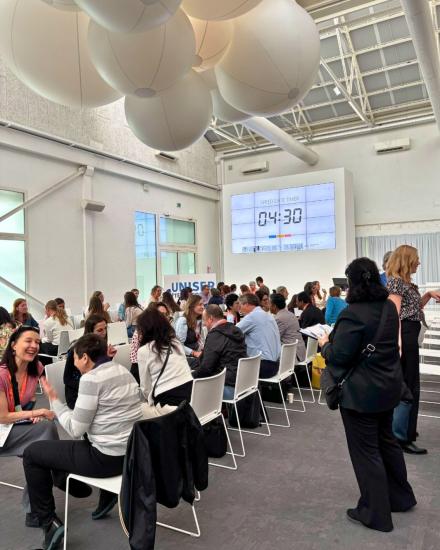 Suuri tila täynnä ihmisiä osallistumassa "speed date" -muotoiseen verkostoitumistilanteeseen. Parit keskustelevat vastakkain istuen, ja taustalla näkyy suuri näyttö, jossa ajastin laskee viiden minuutin keskusteluaikaa. Katossa roikkuu suuria pyöreitä koristeita ja tilassa vallitsee aktiivinen ja iloinen ilmapiiri.