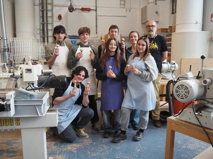 Ryhmä nuoria seisoo puuverstaassa opettajansa kanssa ja poseeraa hymyillen kameralle. Kaikilla on yllään työesiliinat, ja he pitelevät käsissään sorvattuja puuesineitä, kuten pieniä sieniä. Edustalla on sorvikoneita ja lattialla puupölyä. Taustalla näkyy verstaan valkoisia tiiliseiniä, koneita ja puumateriaaleja. Tunnelma on iloinen ja ylpeä kädentaidoista.