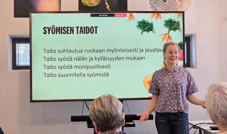Aliisa Hyvönen seisoo esityksen edessä ja puhuu yleisölle. Taustalla on dia, jossa lukee "SYÖMISEN TAIDOT" ja listaus muun muassa taidoista suhtautua ruokaan myönteisesti ja syödä monipuolisesti. Esityksessä käytetään pirteitä värejä ja kasvikuvitusta.