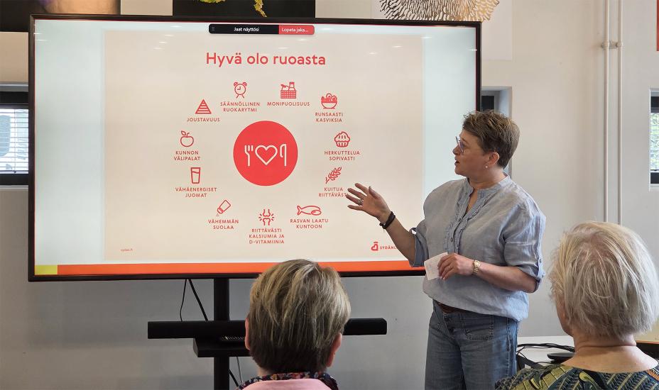 Maria Hietto esittelee yleisölle diaesitystä, jossa keskellä on ruokailuun liittyvä ikoni ja ympärillä eri teemoja, kuten ”Säännöllinen ruokarytmi”, ”Runsaasti kasviksia” ja ”Riittävästi kalsiumia ja D-vitamiinia”. Yleisön joukossa on iäkkäämpiä osallistujia.