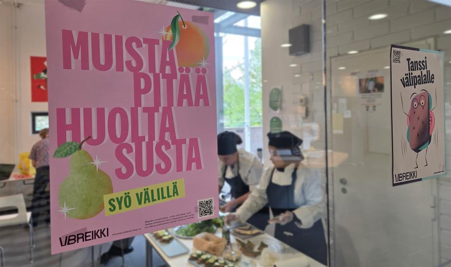 Lasiseinään kiinnitetyssä vaaleanpunaisessa julisteessa lukee isolla “Muista pitää huolta susta – syö välillä” ja siinä on päärynän ja persikan kuvat. Vieressä toinen juliste, jossa lukee “Tanssi välipalalle” ja näkyy piirretty hahmo, jolla on sämpylän näköinen vartalo. Taustalla kaksi kokkiasuista opiskelijaa valmistaa leipiä pöydän ääressä.