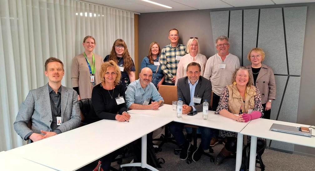 Opetusministeriön kutsumia henkilöitä valkoisen kokouspöydän ääressä. Osa istuu ja osa seisoo.