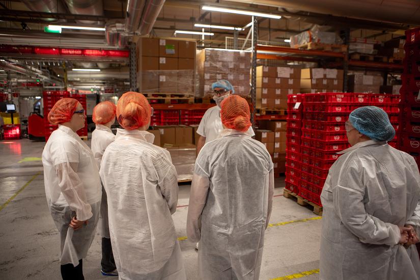 Joukko opiskelijoita seisoo suojavaatteet päällä isossa hallissa. Taustalla näkyy punaisia laatikoita.