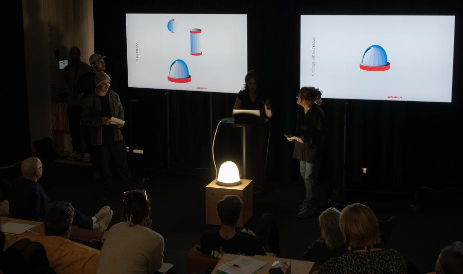 Opiskelijaryhmä esittelee valaisinprototyyppiä yleisölle kahden teknisiä kuvia näyttävän näytön edessä.