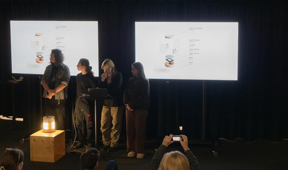 Ryhmä esittelee tuotteen visuaalisia inspiraatiokuvia kahdella näytöllä ja demonstroi prototyyppiä puukuution päällä.