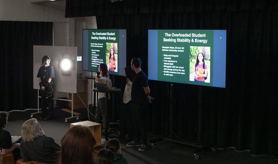 Ryhmä esittelee asiakasprofiilia kahdella näytöllä; vieressä opiskelija seisoo valonäytteen edessä sen vaikutusta havainnollistaen.