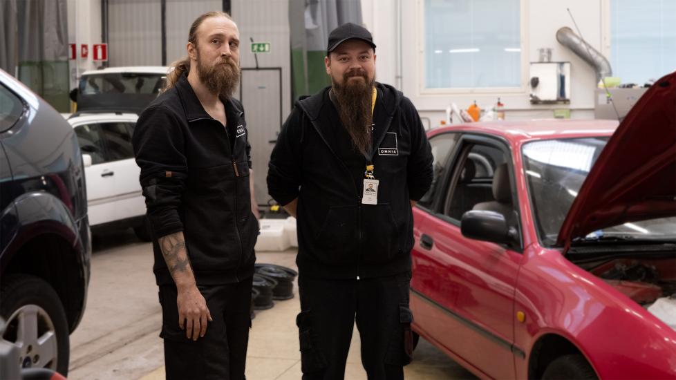 Kaksi henkilöä seisoo autokorjaamossa auton vieressä ja katsoo kameraan.