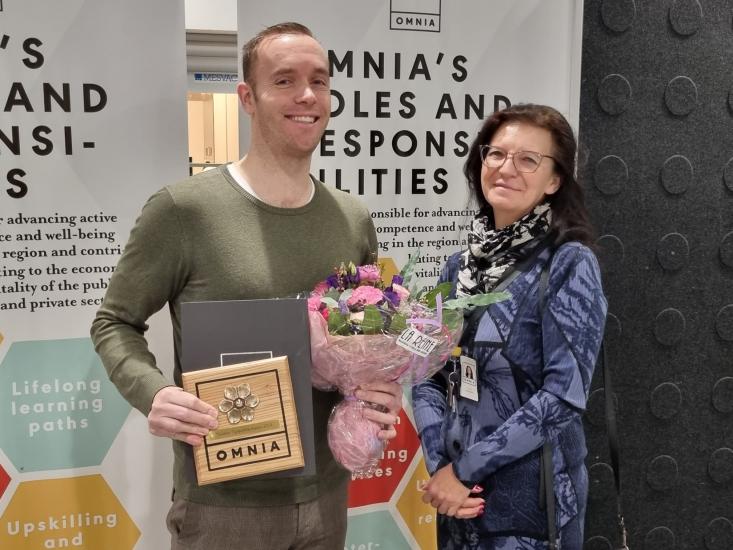 Kaksi henkilöä seisoo Omnian esittelytaustan edessä. Toinen pitää käsissään kukkakimppua ja palkintoa.
