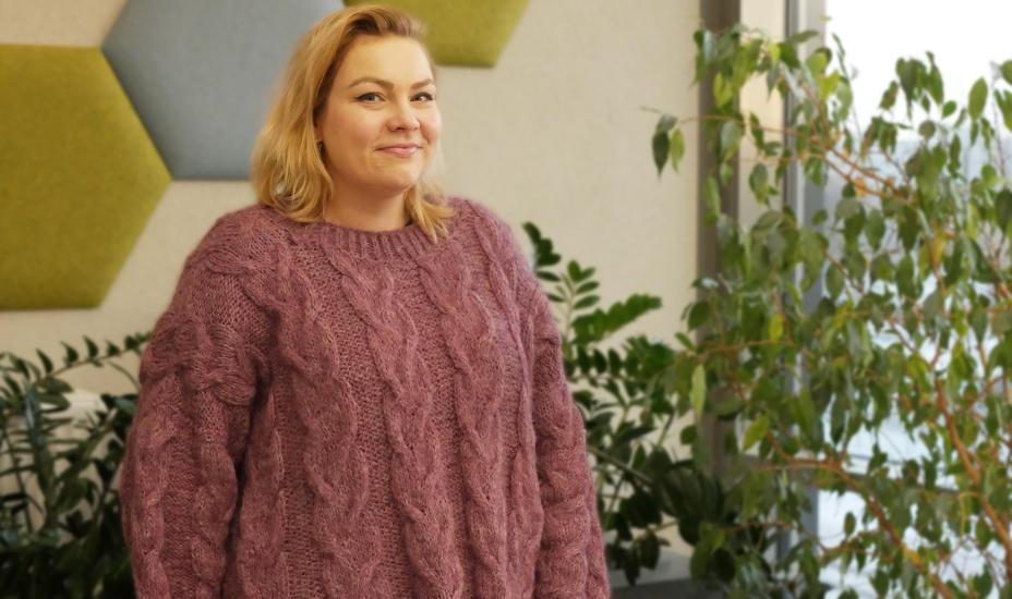 Nainen seisoo sisätilassa kasvien ja akustiikkapaneelien edessä ja hymyilee.