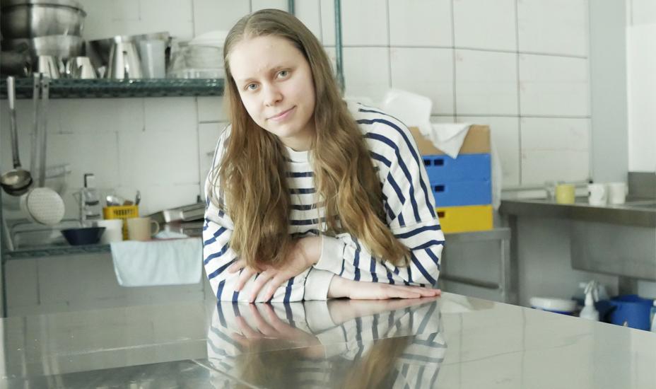 Nuori nainen nojaa pöytään ammattikeittiössä ja katsoo kameraan.