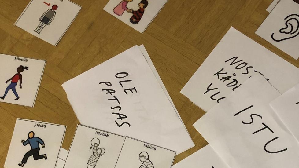 Lattialla on suomen kielen oppimiskortteja, joissa on kuvia ja sanoja liikkumisesta, sekä käsin kirjoitettuja paperilappuja.