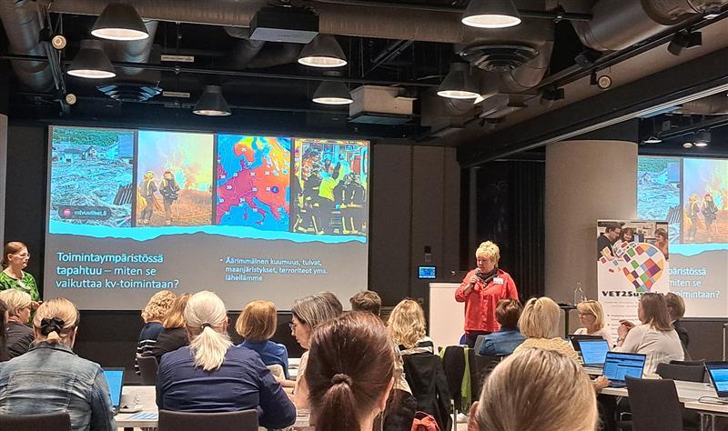 Henkilö pitää esitystä seminaaritilassa, jossa yleisö seuraa luentoa kannettavien tietokoneiden äärellä. Taustalla näkyy esitysdia toimintaympäristön vaikutuksista kansainväliseen toimintaan.