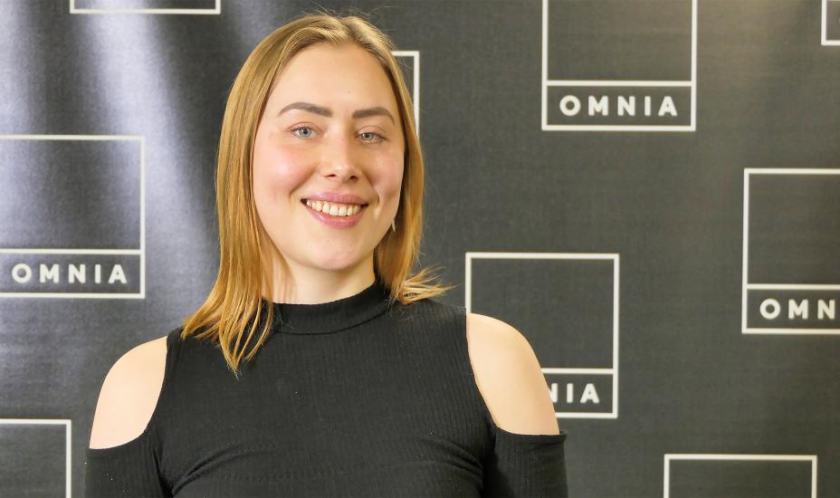 Henkilö hymyilee kameralle Omnian logolla varustetun taustaseinän edessä.