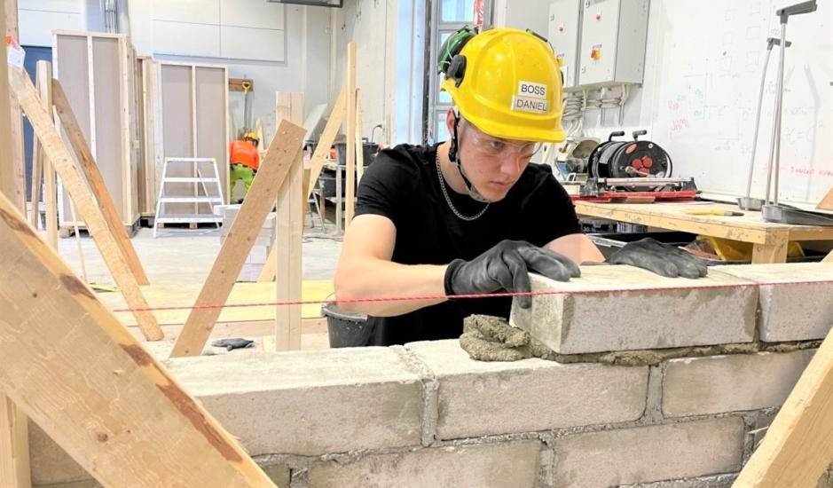 Opiskelija työskentelee rakennusalalla ja muurtaa tiiliseinää suojakypärä päässä sisätiloissa.