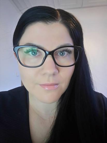 Lähikuva henkilöstä, jolla on pitkät tummat hiukset ja silmälasit, katsoo suoraan kameraan.